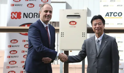 Two men in suits shake hands on a deck, a heater between them, with company banners in the background
